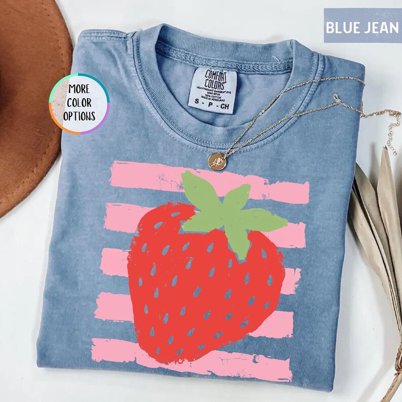 May include: A blue jean colored t-shirt featuring a large red strawberry graphic with green leaves centered over horizontal light pink brushstroke stripes. The shirt has a crew neckline and a Comfort Colors brand label at the inner collar.