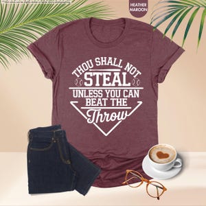 May include: A heather maroon t-shirt with the text "THOU SHALL NOT STEAL UNLESS YOU CAN BEAT THE THROW" in white. The shirt is paired with dark blue jeans, a cup of coffee with a heart design, and eyeglasses.