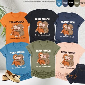 May include: A collection of t-shirts in various colors, each featuring a cartoon monkey hugging another monkey. The text "TEAM PUNCH" and "You are never alone!" are printed on each shirt. A pair of jeans, shoes, and glasses are also in the image.