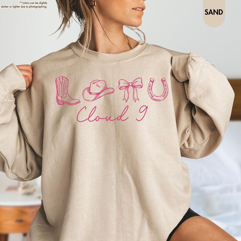 May include: A sand-coloured sweatshirt featuring a pink graphic of a cowboy boot, hat, bow, and horseshoe, with the text "Cloud 9". The sweatshirt has a crew neck and is a neutral colour.