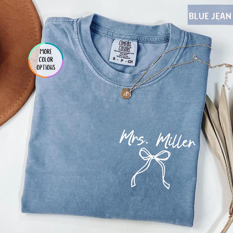 Puede incluir: Una camiseta azul vaquero con el texto "Mrs. Miller" y un lazo blanco. Un collar dorado con un colgante est&aacute; sobre la camiseta. La camiseta est&aacute; doblada y colocada sobre una superficie blanca.