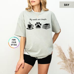 May include: A light green t-shirt with the phrase "My needs are simple." and illustrations of a coffee cup, paw print, and books. The shirt is a relaxed fit and has short sleeves.