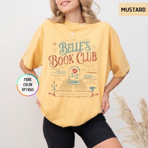 Pu&ograve; includere: T-shirt color senape con la scritta "Belle's Book Club" in un design vintage. La grafica include una rosa in una cupola di vetro, un libro aperto e la frase "Tale as Old as Time".