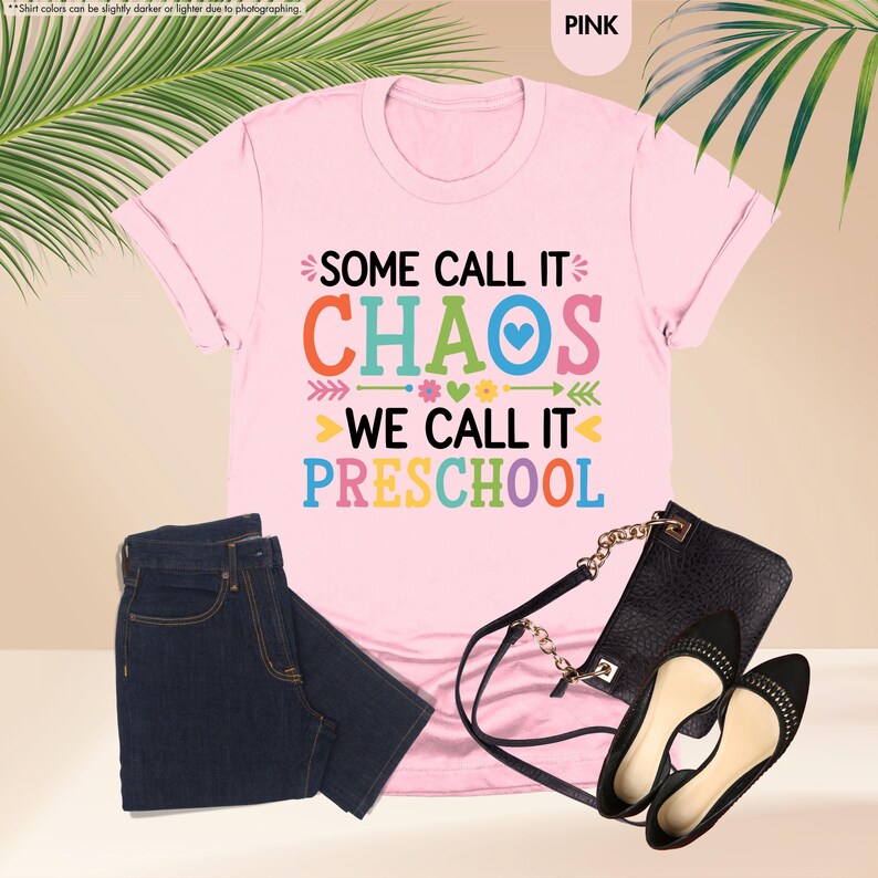 May include: Pink t-shirt with the text "SOME CALL IT CHAOS WE CALL IT PRESCHOOL" in colorful letters. The shirt is paired with dark blue jeans, a black purse, and black flats.