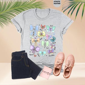 May include: A light grey t-shirt with colourful bow illustrations and character names such as Cinderella and Ariel. The image also shows dark blue jeans, pink trainers, and a small pink wallet.