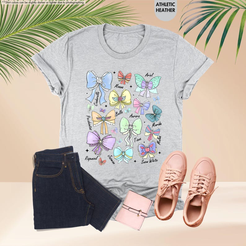 May include: A light grey t-shirt with colourful bow illustrations and character names such as Cinderella and Ariel. The image also shows dark blue jeans, pink trainers, and a small pink wallet.