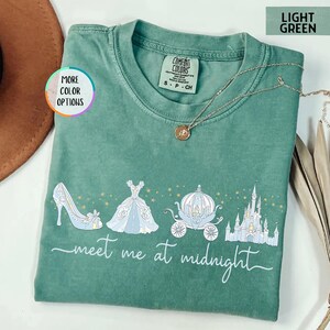 May include: Light green t-shirt with a Cinderella-themed graphic. The design includes a glass slipper, ball gown, carriage, and castle, with the text "meet me at midnight". The shirt is a Comfort Colors brand.