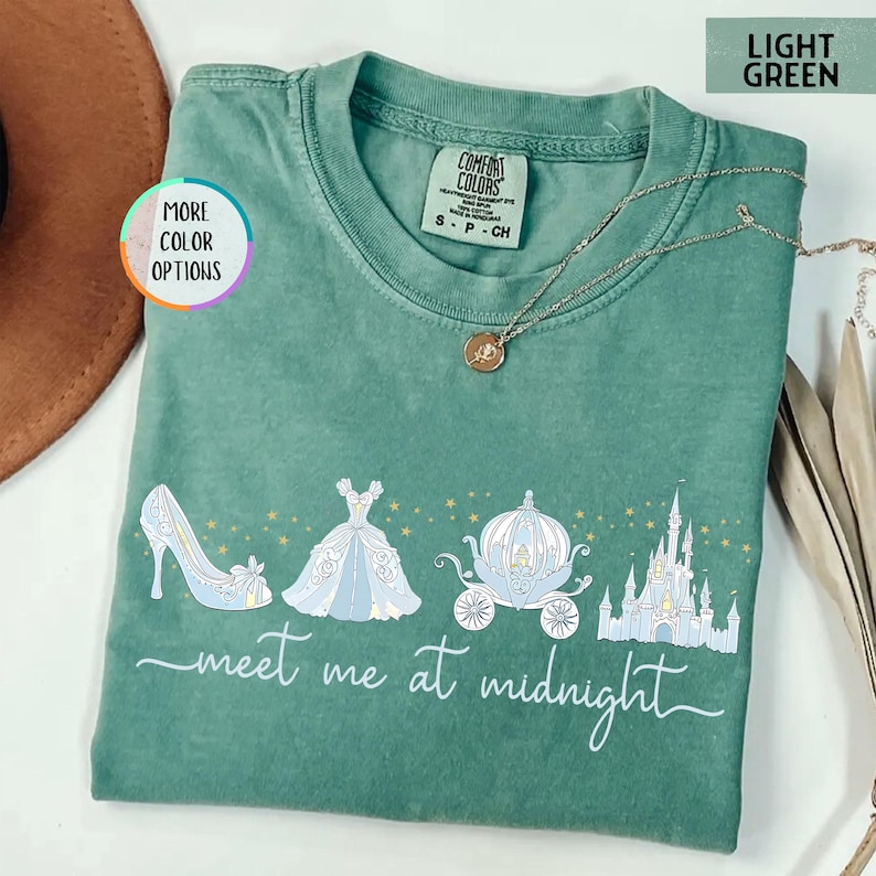 May include: Light green t-shirt with a Cinderella-themed graphic. The design includes a glass slipper, ball gown, carriage, and castle, with the text "meet me at midnight". The shirt is a Comfort Colors brand.