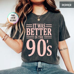 May include: A charcoal gray short-sleeve t-shirt featuring the light pink text "IT WAS BETTER IN THE 90's" printed on the front. The shirt has a crew neckline and a vintage-style washed finish. A small circular badge reads "MORE COLOR OPTIONS".