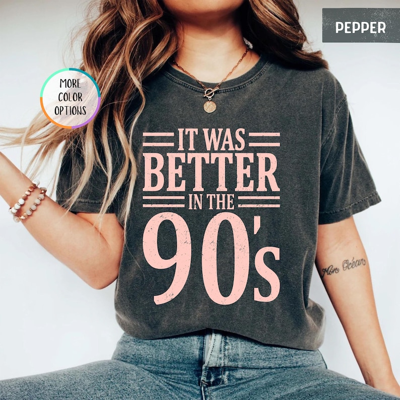 May include: A charcoal gray short-sleeve t-shirt featuring the light pink text "IT WAS BETTER IN THE 90's" printed on the front. The shirt has a crew neckline and a vintage-style washed finish. A small circular badge reads "MORE COLOR OPTIONS".
