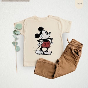 May include: A cream-colored t-shirt featuring a classic Mickey Mouse graphic in black, red, and white. Paired with brown pants. The image has a natural aesthetic.