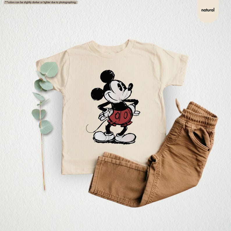 May include: A cream-colored t-shirt featuring a classic Mickey Mouse graphic in black, red, and white. Paired with brown pants. The image has a natural aesthetic.