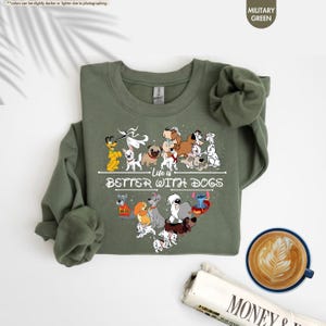 May include: Military green sweatshirt with a heart-shaped design of various dog breeds and the text "Life is Better With Dogs." The sweatshirt has long sleeves and a crew neck. A cup of coffee and a newspaper are in the background.