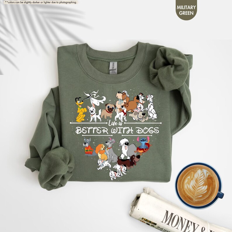 May include: Military green sweatshirt with a heart-shaped design of various dog breeds and the text "Life is Better With Dogs." The sweatshirt has long sleeves and a crew neck. A cup of coffee and a newspaper are in the background.