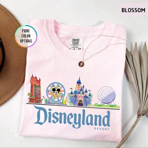 Op de afbeelding: Lichtroze t-shirt met een Disneyland Resort afbeelding. De afbeelding toont iconische parklandmarks en het Disneyland-logo in blauw. Het shirt heeft een ronde hals en een Comfort Colors label.