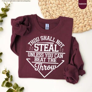 May include: Maroon sweatshirt with the text "THOU SHALL NOT STEAL UNLESS YOU CAN BEAT THE THROW" in white. The design includes baseballs and a baseball diamond shape. The sweatshirt is a crew neck.