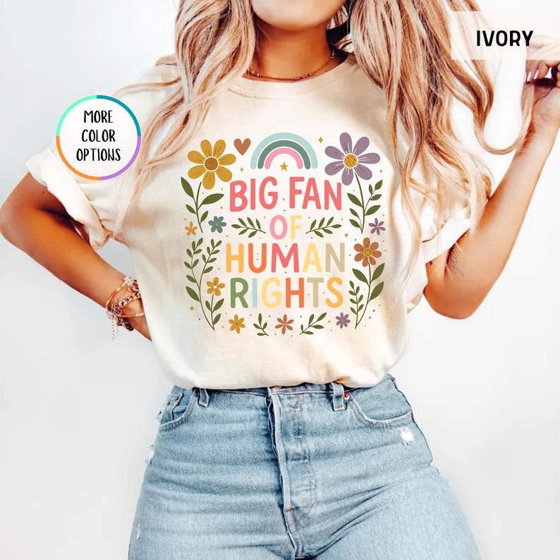 May include: Ivory-colored t-shirt with the text "BIG FAN OF HUMAN RIGHTS" in colorful letters. The design includes a rainbow, flowers, and leaves. The shirt is a crew neck and has short sleeves.