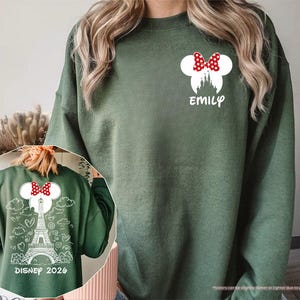 May include: A military green sweatshirt featuring a white graphic on the left chest of a castle silhouette with a red and white polka dot bow and the name EMILY. The back displays a white line drawing of the Eiffel Tower, clouds, and hearts.