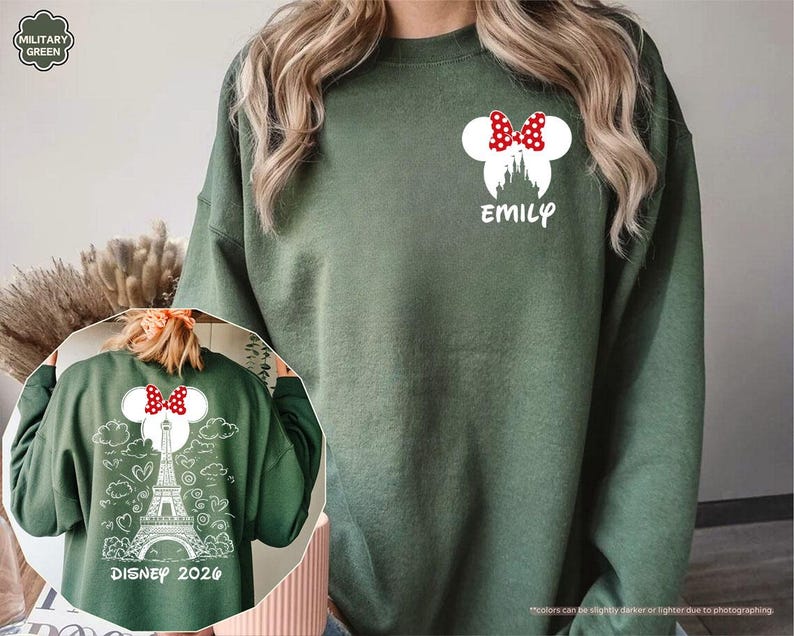 May include: A military green sweatshirt featuring a white graphic on the left chest of a castle silhouette with a red and white polka dot bow and the name EMILY. The back displays a white line drawing of the Eiffel Tower, clouds, and hearts.