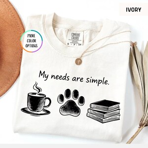 May include: Ivory-colored t-shirt with the phrase "My needs are simple." The shirt features black illustrations of a coffee cup, a paw print, and a stack of books. The shirt has a crew neck and short sleeves.