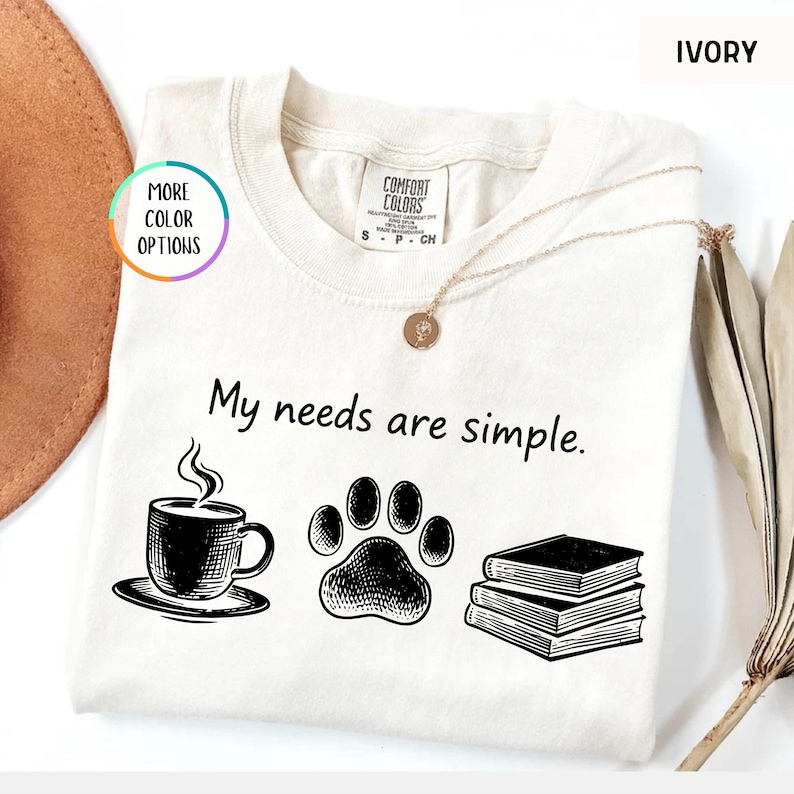 May include: Ivory-colored t-shirt with the phrase "My needs are simple." The shirt features black illustrations of a coffee cup, a paw print, and a stack of books. The shirt has a crew neck and short sleeves.