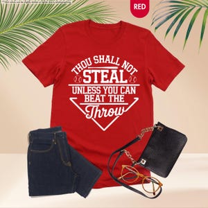 May include: A red t-shirt with the text "THOU SHALL NOT STEAL UNLESS YOU CAN BEAT THE THROW" in white. The image also shows a pair of dark blue jeans, a black purse, and eyeglasses.