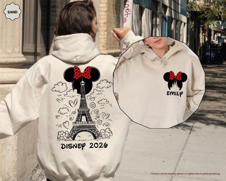 May include: A sand-colored hooded sweatshirt featuring a black graphic of the Eiffel Tower topped with a Minnie Mouse silhouette and red polka-dot bow. The back includes clouds, hearts, and the text DISNEY 2026. The front chest shows the same icon with the name EMILY.