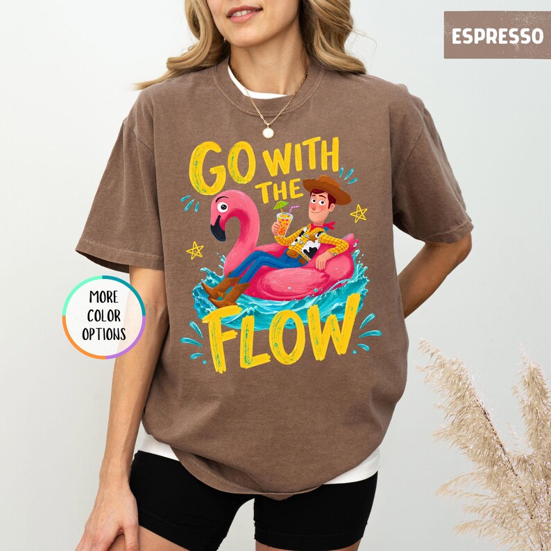 Puede incluir: Una camiseta color espresso con un gr&aacute;fico de un personaje vaquero de dibujos animados flotando en un flotador de flamenco rosa en agua azul. El texto dice "GO WITH THE FLOW" en letras amarillas. Un peque&ntilde;o c&iacute;rculo gr&aacute;fico dice "MORE COLOR OPTIONS".