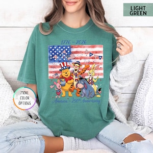 May include: Light green t-shirt featuring a graphic of Winnie the Pooh and friends in front of an American flag. The graphic includes the text "1776-2026" and "America - 250th Anniversary".