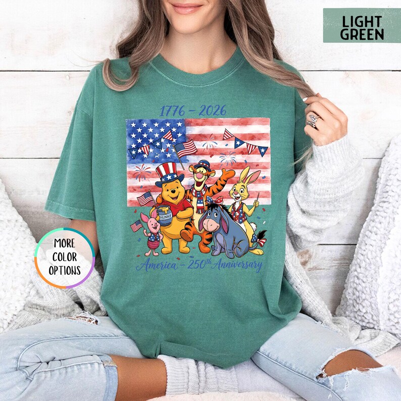 May include: Light green t-shirt featuring a graphic of Winnie the Pooh and friends in front of an American flag. The graphic includes the text "1776-2026" and "America - 250th Anniversary".