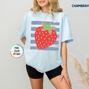 May include: A light blue short-sleeve t-shirt featuring a large red strawberry graphic centered over horizontal grey stripes. The shirt includes the text CHAMBRAY in the upper right corner and a circular badge stating MORE COLOR OPTIONS.