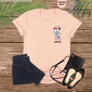 May include: Heather peach t-shirt featuring a Minnie Mouse graphic with the name "Sophie" below. The image includes dark blue jeans, a black purse, and black flats. The shirt's color is described as heather peach.