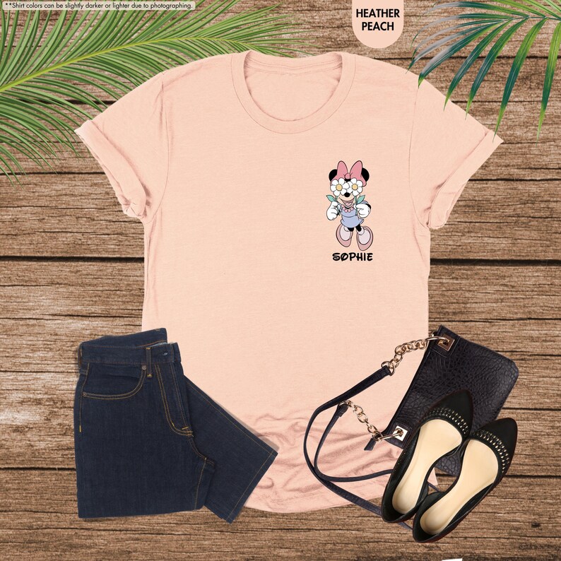 May include: Heather peach t-shirt featuring a Minnie Mouse graphic with the name "Sophie" below. The image includes dark blue jeans, a black purse, and black flats. The shirt's color is described as heather peach.