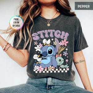 May include: Dark gray t-shirt featuring the word "STITCH" in a retro font, with a cartoon character and floral accents. The design includes a blue alien-like creature, ducks, flowers, and a checkered border.