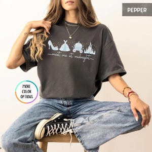 May include: Pepper-colored t-shirt with a light blue graphic of a high heel, dress, carriage, and castle. The text "meet me at midnight" is written below the graphic. The shirt is worn with blue jeans and black sneakers.