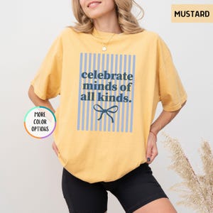 Puede incluir: Camiseta amarilla mostaza con el texto azul "celebrate minds of all kinds". El texto est&aacute; centrado dentro de un rect&aacute;ngulo a rayas azul claro, con un lazo azul en la parte inferior. La camiseta es de estilo casual.