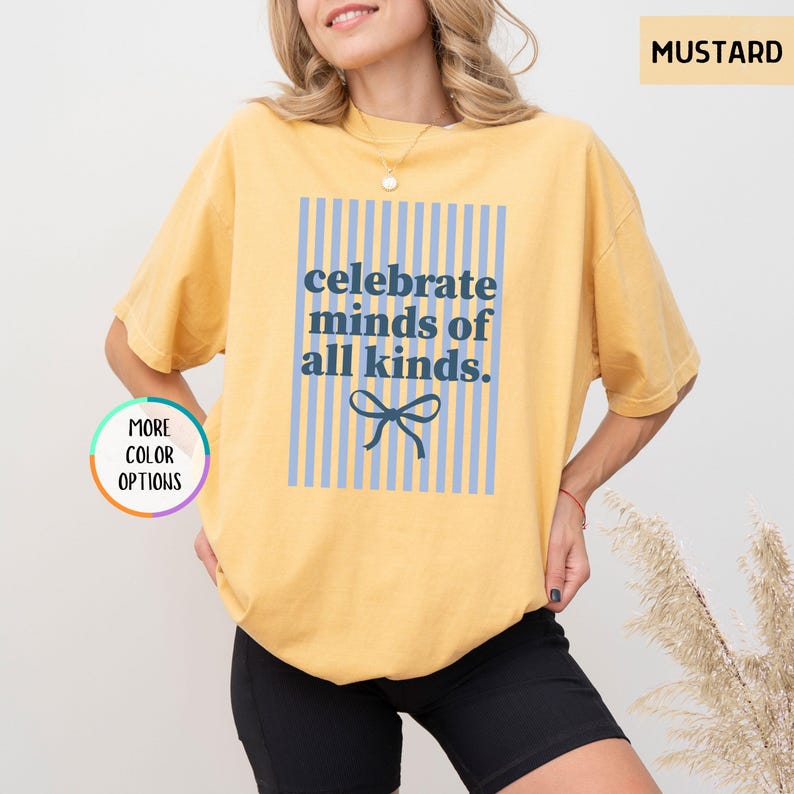 Puede incluir: Camiseta amarilla mostaza con el texto azul "celebrate minds of all kinds". El texto est&aacute; centrado dentro de un rect&aacute;ngulo a rayas azul claro, con un lazo azul en la parte inferior. La camiseta es de estilo casual.