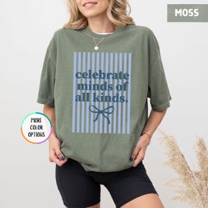 Puede incluir: Camiseta verde musgo con el texto "celebrate minds of all kinds" en azul. El texto est&aacute; dentro de un rect&aacute;ngulo a rayas azules con un lazo azul en la parte inferior. La camiseta es de corte holgado.