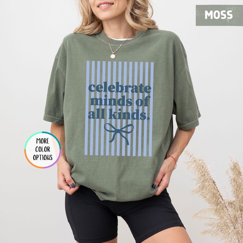 Puede incluir: Camiseta verde musgo con el texto "celebrate minds of all kinds" en azul. El texto est&aacute; dentro de un rect&aacute;ngulo a rayas azules con un lazo azul en la parte inferior. La camiseta es de corte holgado.