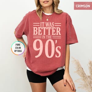May include: A crimson short-sleeved t-shirt with a crew neckline. The front features light pink text that reads IT WAS BETTER IN THE 90's. A small circular graphic on the left side displays the text MORE COLOR OPTIONS. The fabric has a washed look.