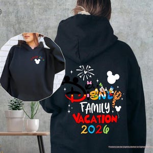 Puede incluir: Una sudadera con capucha negra que presenta un gr&aacute;fico en la parte posterior con el texto Disney Family Vacation 2026, personajes de dibujos animados coloridos y fuegos artificiales. Una versi&oacute;n m&aacute;s peque&ntilde;a est&aacute; en el pecho izquierdo.