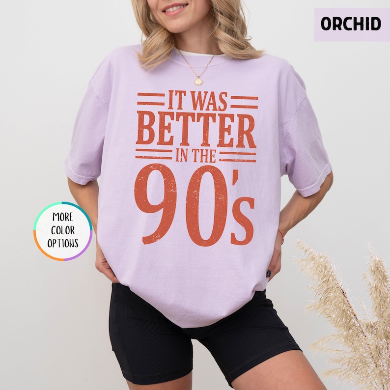 May include: A light purple short-sleeve t-shirt featuring the text IT WAS BETTER IN THE 90's in a distressed orange font. The shirt has a crew neckline and a relaxed fit. A small circular graphic in the corner displays the text MORE COLOR OPTIONS.