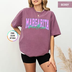 May include: Berry-colored t-shirt with the text "Margarita Weather" in a bold, two-tone design. The shirt has a relaxed fit and short sleeves. The model is wearing black shorts.