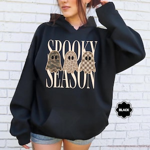 May include: A black hooded sweatshirt featuring the text SPOOKY SEASON in a cream-colored serif font. Below the text are three ghost illustrations with different patterns: one in camouflage, one in leopard print, and one in a brown checkerboard pattern.