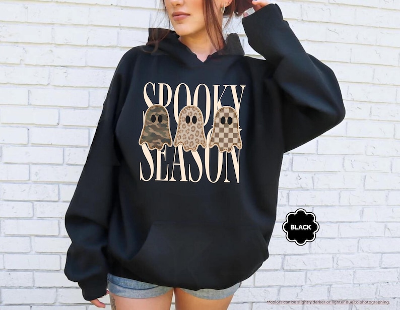 May include: A black hooded sweatshirt featuring the text SPOOKY SEASON in a cream-colored serif font. Below the text are three ghost illustrations with different patterns: one in camouflage, one in leopard print, and one in a brown checkerboard pattern.