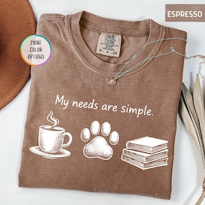 May include: Espresso-colored t-shirt with the phrase "My needs are simple" and illustrations of a coffee cup, paw print, and books. The shirt has a crew neck and a Comfort Colors tag.