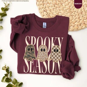 May include: A maroon long-sleeve sweatshirt featuring the text SPOOKY SEASON in large cream letters. Three ghost-shaped graphics are centered, each with a different pattern: one in camouflage, one in leopard print, and one in a brown checkerboard.