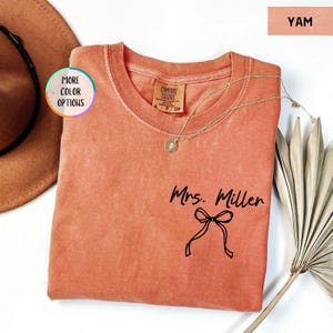 Puede incluir: Una camiseta Comfort Colors color &ntilde;ame con el nombre "Mrs. Miller" y un gr&aacute;fico de lazo impreso en negro. Un collar de oro con un colgante descansa sobre la camisa. La camisa se muestra con un sombrero y una planta seca.