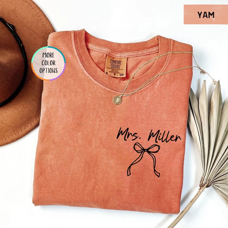Puede incluir: Una camiseta Comfort Colors color &ntilde;ame con el nombre "Mrs. Miller" y un gr&aacute;fico de lazo impreso en negro. Un collar de oro con un colgante descansa sobre la camisa. La camisa se muestra con un sombrero y una planta seca.