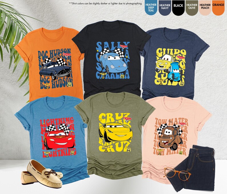 以下が含まれることがあります： 様々な色の半袖Tシャツのコレクションで、それぞれに漫画の車のデザインとテキストが描かれています。シャツの色はオレンジ、黒、ネイビー、オリーブ、ピーチ、ティールです。デザインには、映画「カーズ」のキャラクターとテキストが含まれています。